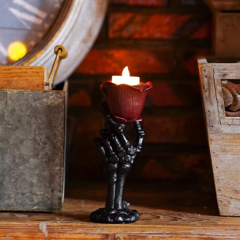 A skeleton hand candle holder holding a dark red lotus-shaped candle holder with a lit white candle, set on a rustic wooden surface with a warm, dimly lit background.