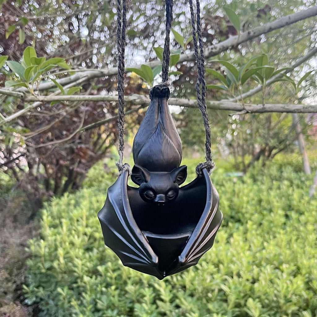 A black hanging bat-shaped ornament or planter suspended by braided cords from tree branches outdoors, with green foliage blurred in the background.