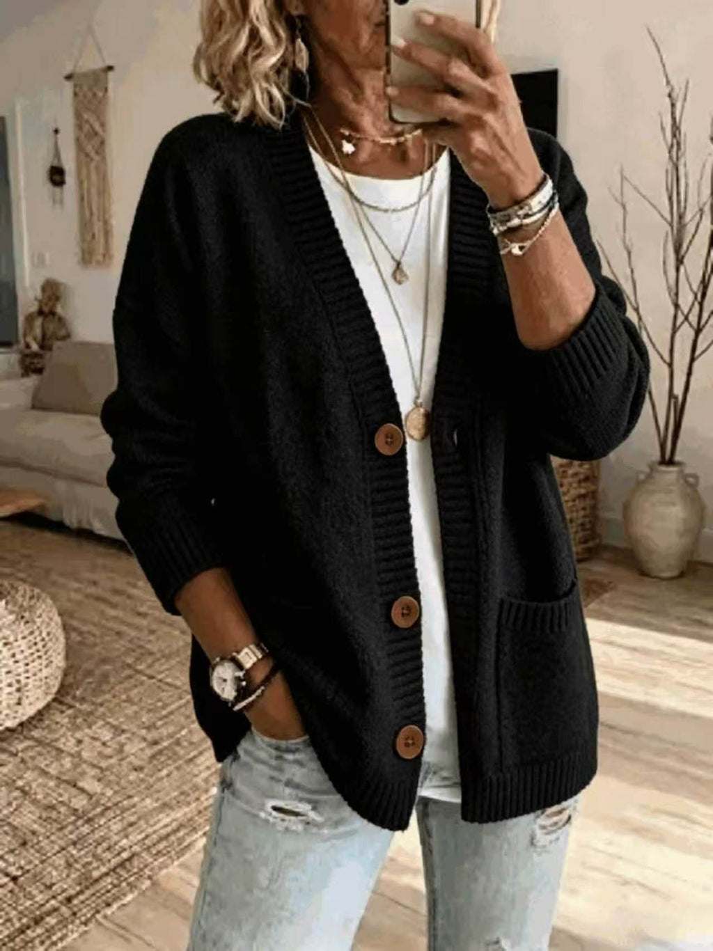 A woman takes a mirror selfie wearing a black knit cardigan with brown buttons over a white top and light-wash distressed jeans, accessorized with layered necklaces, bracelets, and a watch.