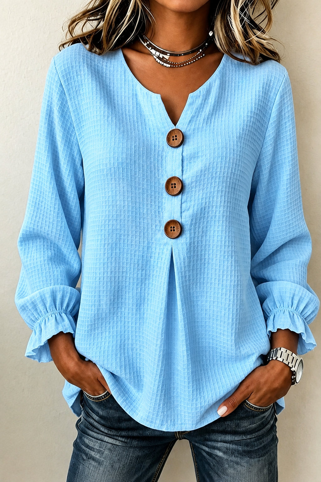 A woman wearing a light blue waffle-textured blouse with a split neckline, three brown buttons, and ruffled cuffs, paired with dark jeans.