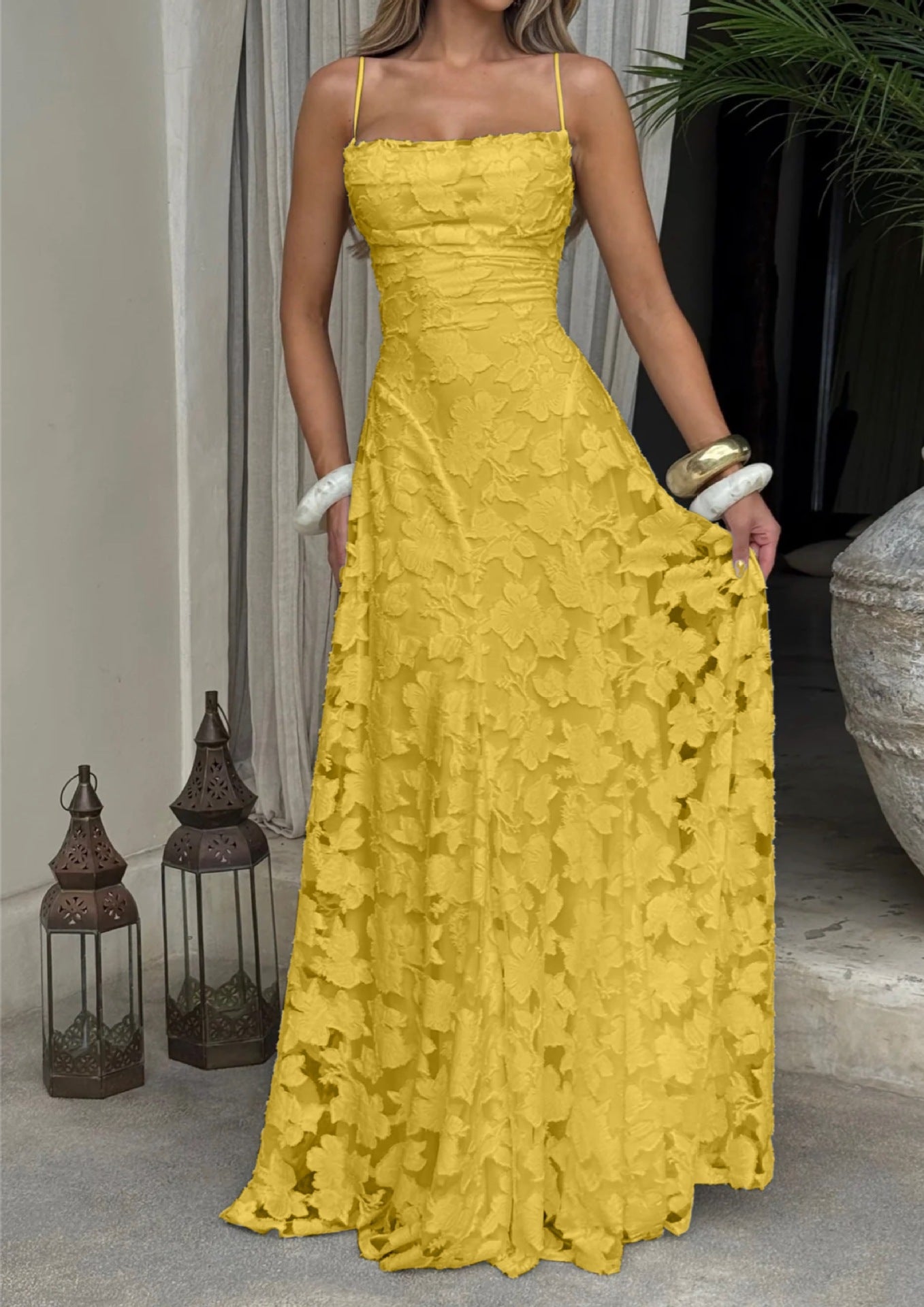 Woman wearing a yellow lace dress standing indoors with decorative lanterns and plants in the background.