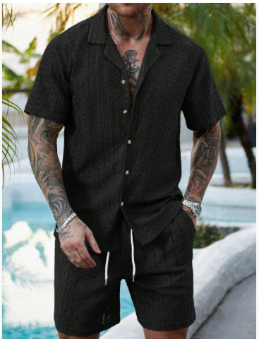 A man wearing a black short-sleeve button-up shirt and matching black drawstring shorts, photographed outdoors near a pool with palm trees in the background.