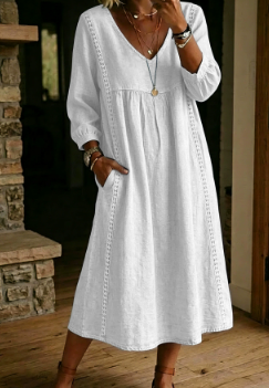 A woman wearing a loose white midi dress with long sleeves, a V-neckline, and side pockets, standing in a rustic indoor setting with a stone wall and wooden floor.