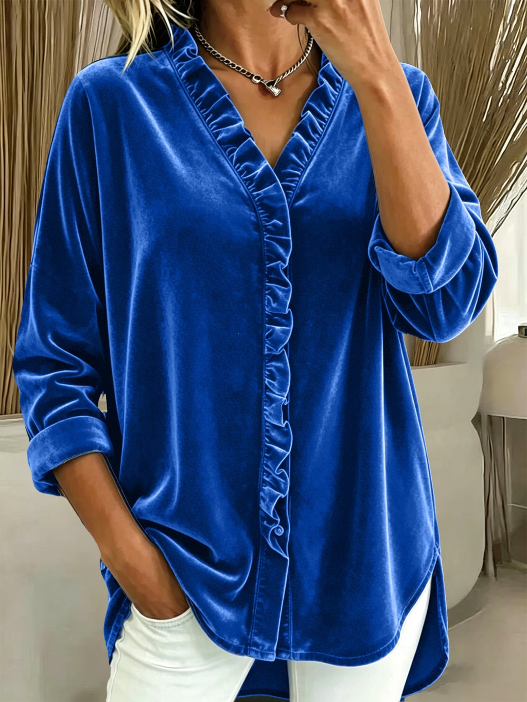 A woman wearing a royal blue velvet long-sleeve blouse with ruffle trim along the V-neck and front placket, paired with white pants in an indoor setting.