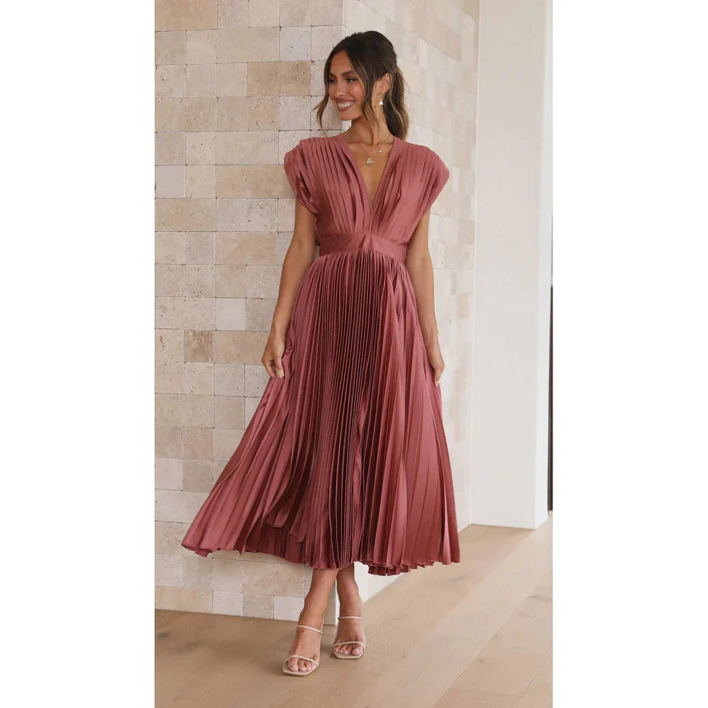 Woman wearing a pink pleated dress standing against a light-colored wall.