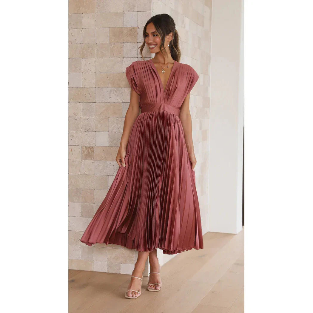 Woman wearing a pink pleated dress standing against a light-colored wall.