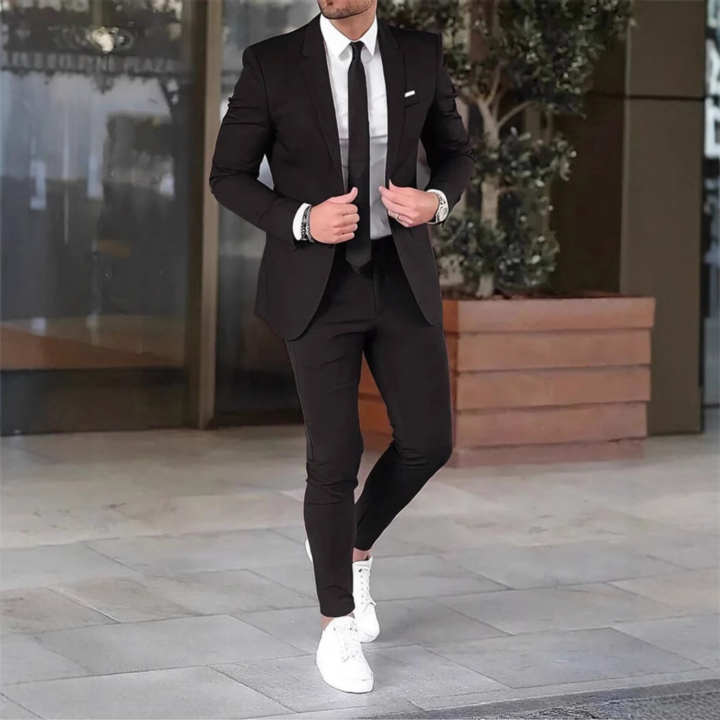 Man in a black suit with white shirt and white sneakers standing on a pavement.