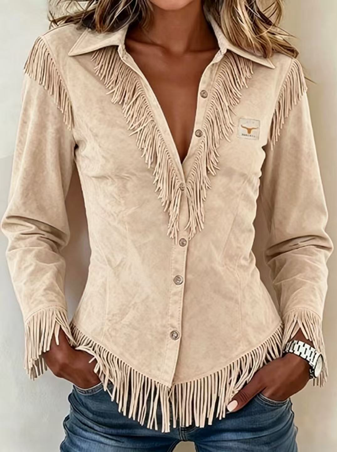 A woman wearing a beige suede long-sleeve western-style shirt with fringe trim and a small embroidered logo patch on the chest, paired with blue jeans.