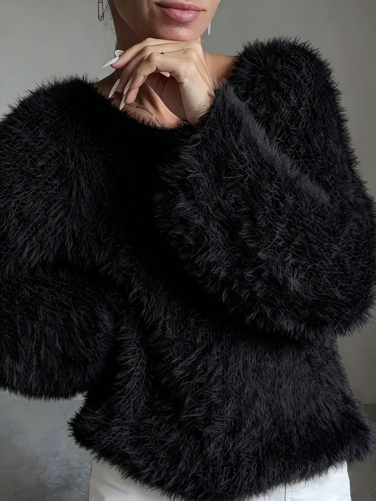 A close-up of a person wearing a voluminous black faux fur sweater with wide, balloon sleeves, paired with white pants against a neutral gray background.