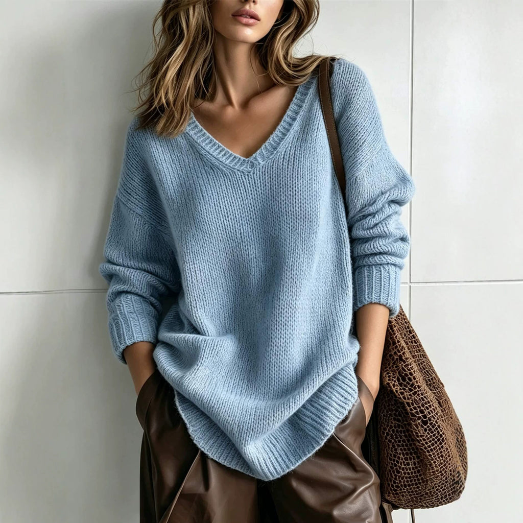 A woman wearing an oversized light blue knit V-neck sweater, styled with brown leather pants and a brown woven shoulder bag, standing against a light gray tiled wall.