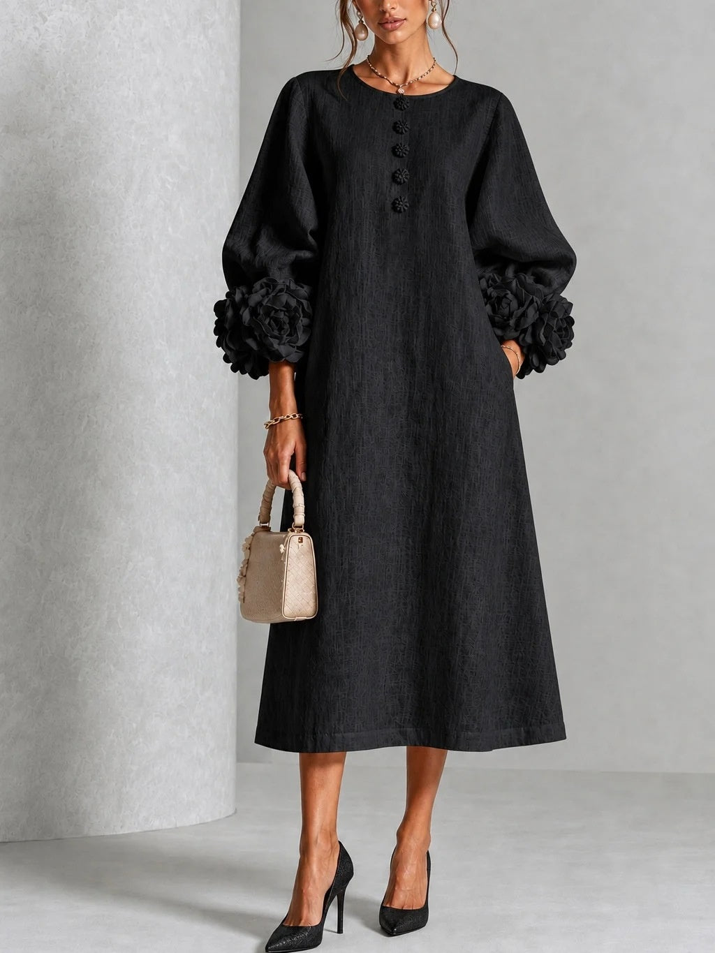 A woman models a black midi dress with voluminous long sleeves, ruffled trim at the cuffs, and a buttoned round neckline. She is holding a small beige handbag and wearing black high heels against a neutral gray studio background.