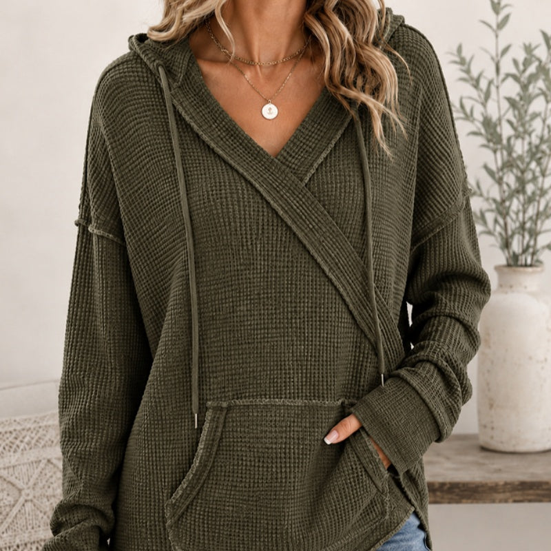 A woman wearing an olive green waffle-knit hooded sweater with a wrap-style V-neck and front pockets, shown in a softly styled indoor setting with a vase and plant in the background.
