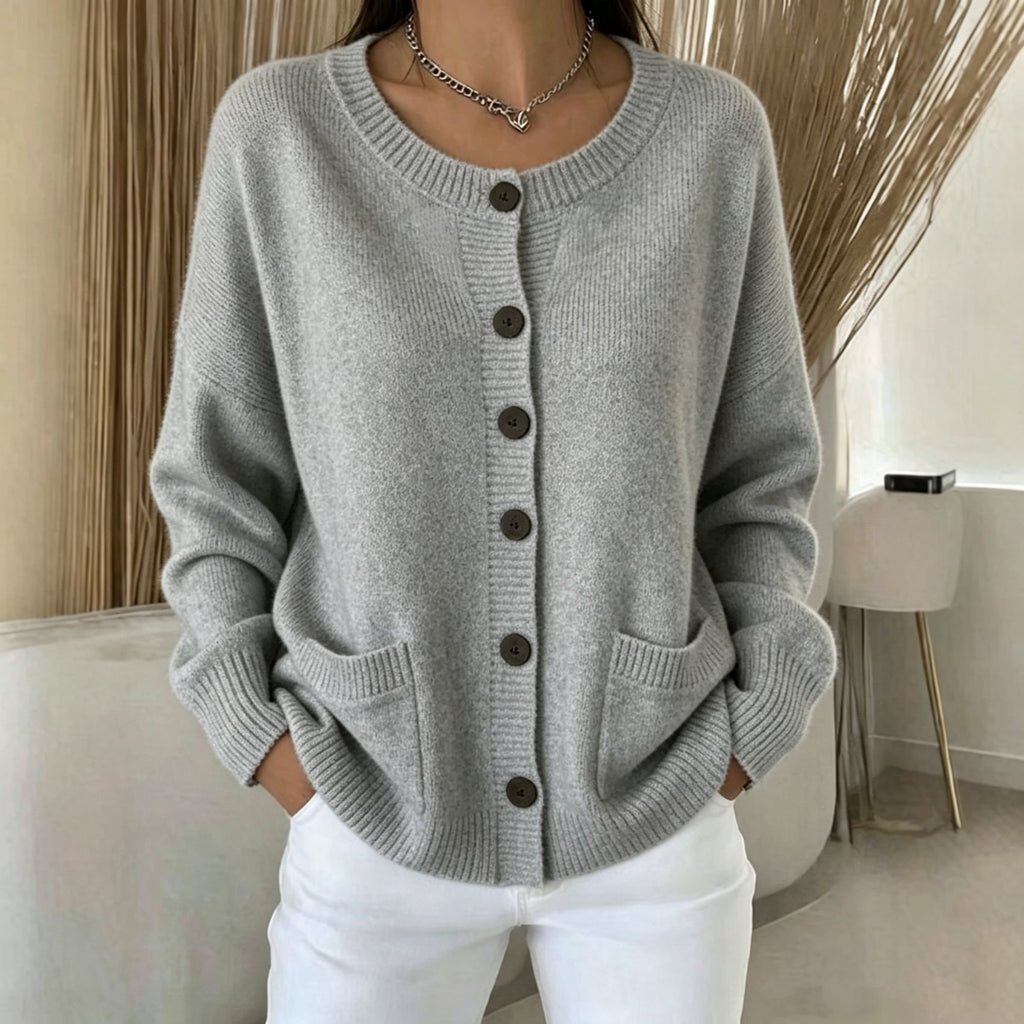 A woman wearing a light gray knit cardigan sweater with dark buttons and white pants, standing in a modern indoor setting with neutral decor.