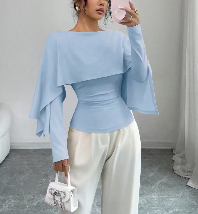 A woman modeling a light blue asymmetrical long-sleeve top with draped side panels and a fitted waist, paired with white pants, holding a small white handbag in a bright indoor room.