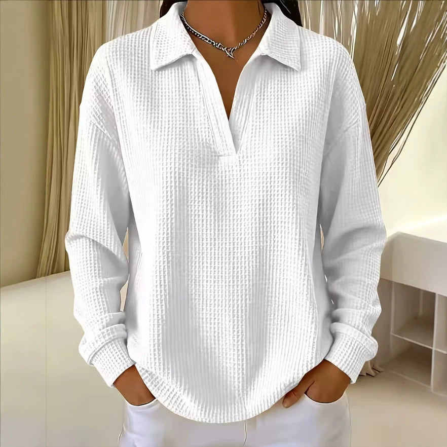 A woman wearing a white long-sleeve waffle-knit polo shirt with a split collar and split placket, paired with white pants and a silver chain necklace, standing in an indoor setting with beige curtains.