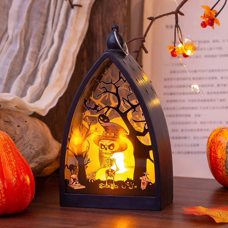 A Halloween-themed decorative lantern with a black arched frame, featuring a skeleton wearing a witch hat in a spooky tree scene with pumpkins and warm yellow backlighting, displayed on a wooden surface with seasonal props.