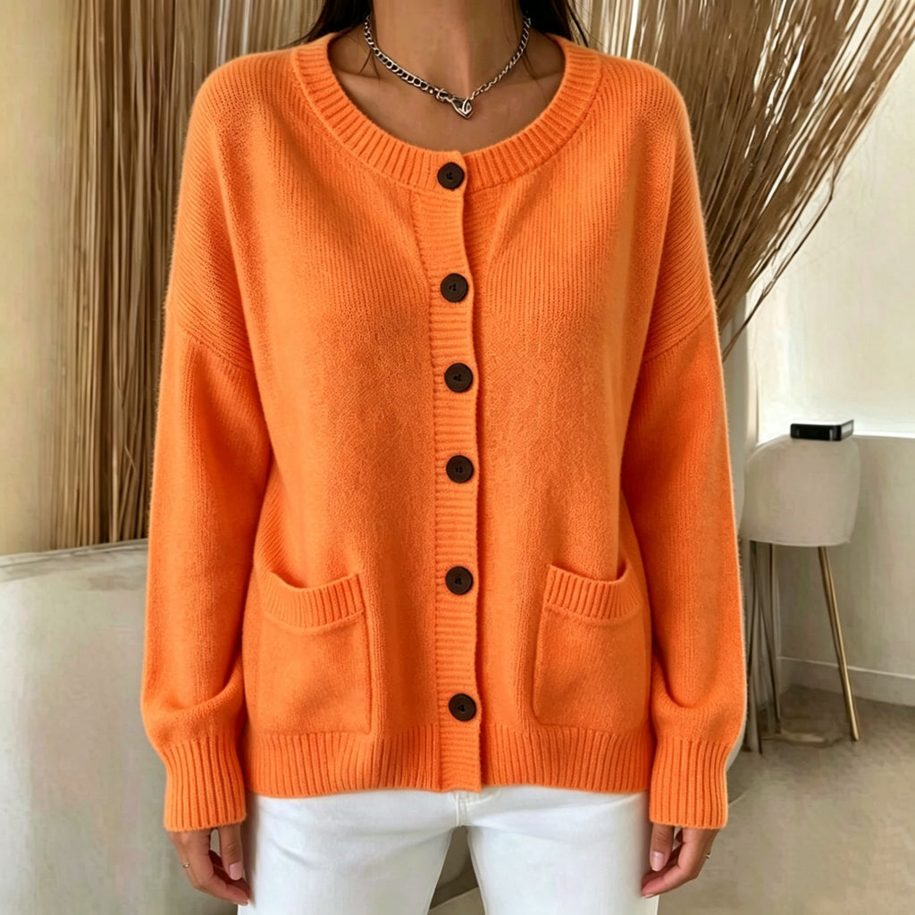 A woman wearing a bright orange knit cardigan sweater with dark brown buttons and front pockets, paired with white pants, in an indoor setting with neutral decor.