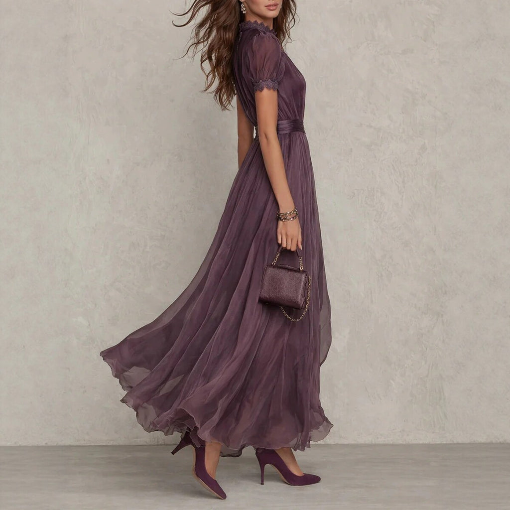 Woman wearing a long purple dress holding a matching handbag against a neutral background