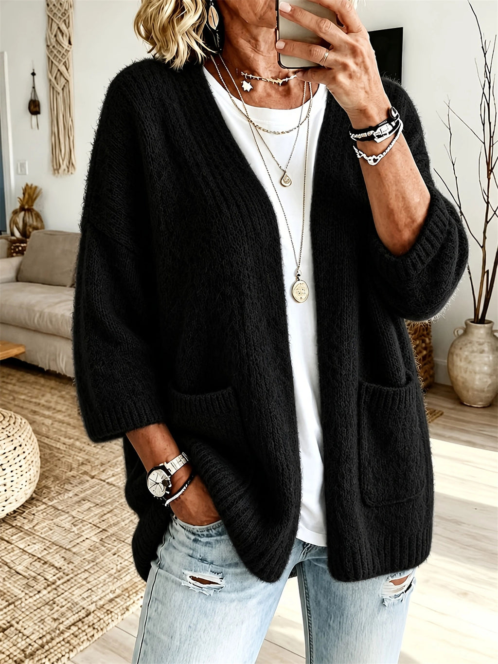 A woman wearing a black open-front cable-knit cardigan with large front pockets, layered over a white t-shirt and light-wash distressed jeans, in a cozy living room setting.
