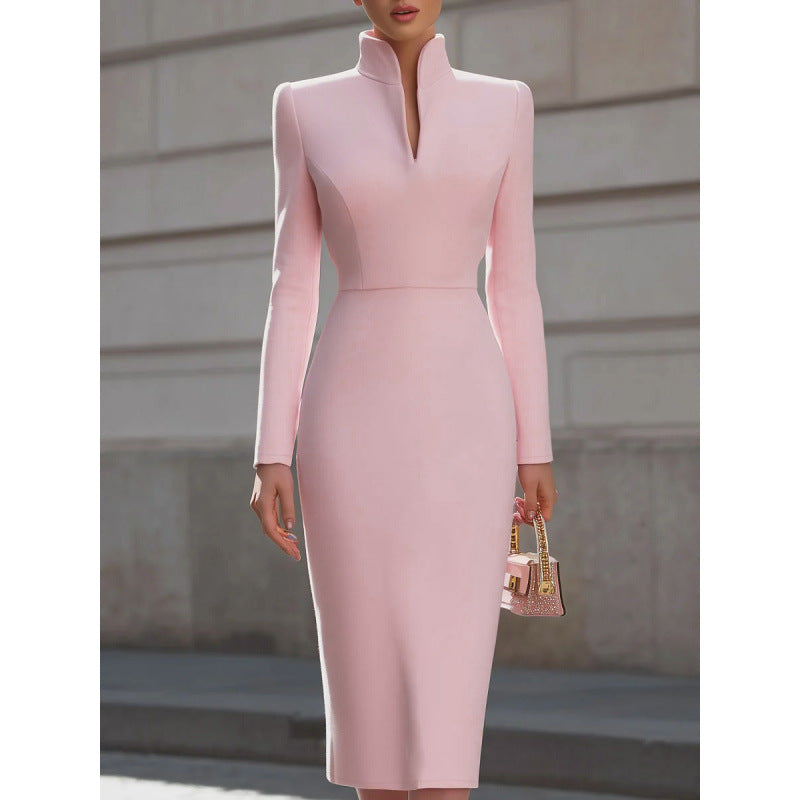 A woman wearing a fitted light pink long-sleeve midi dress with a high mock neck and front slit, holding a small pink handbag, photographed outdoors against a light stone wall.