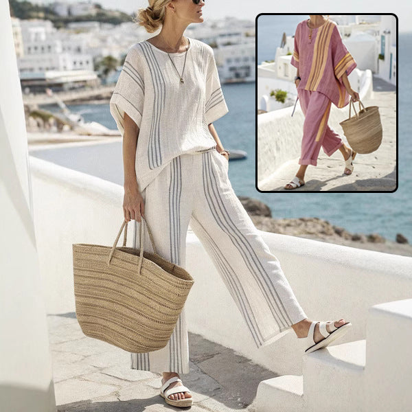 Woman in a white outfit with striped pants holding a straw bag, standing on a balcony with ocean view.