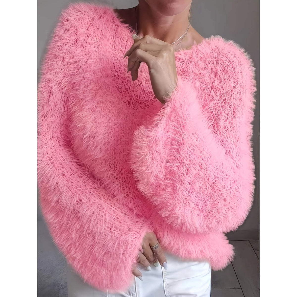 A woman wearing a bright pink, long-pile fuzzy knit sweater with a wide crew neckline and voluminous sleeves, paired with white pants.