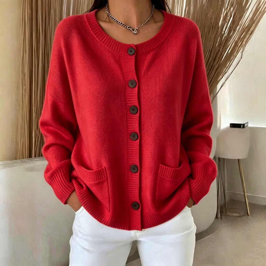 A woman wearing a bright red knit cardigan sweater with dark buttons and front pockets, paired with white pants, standing indoors.