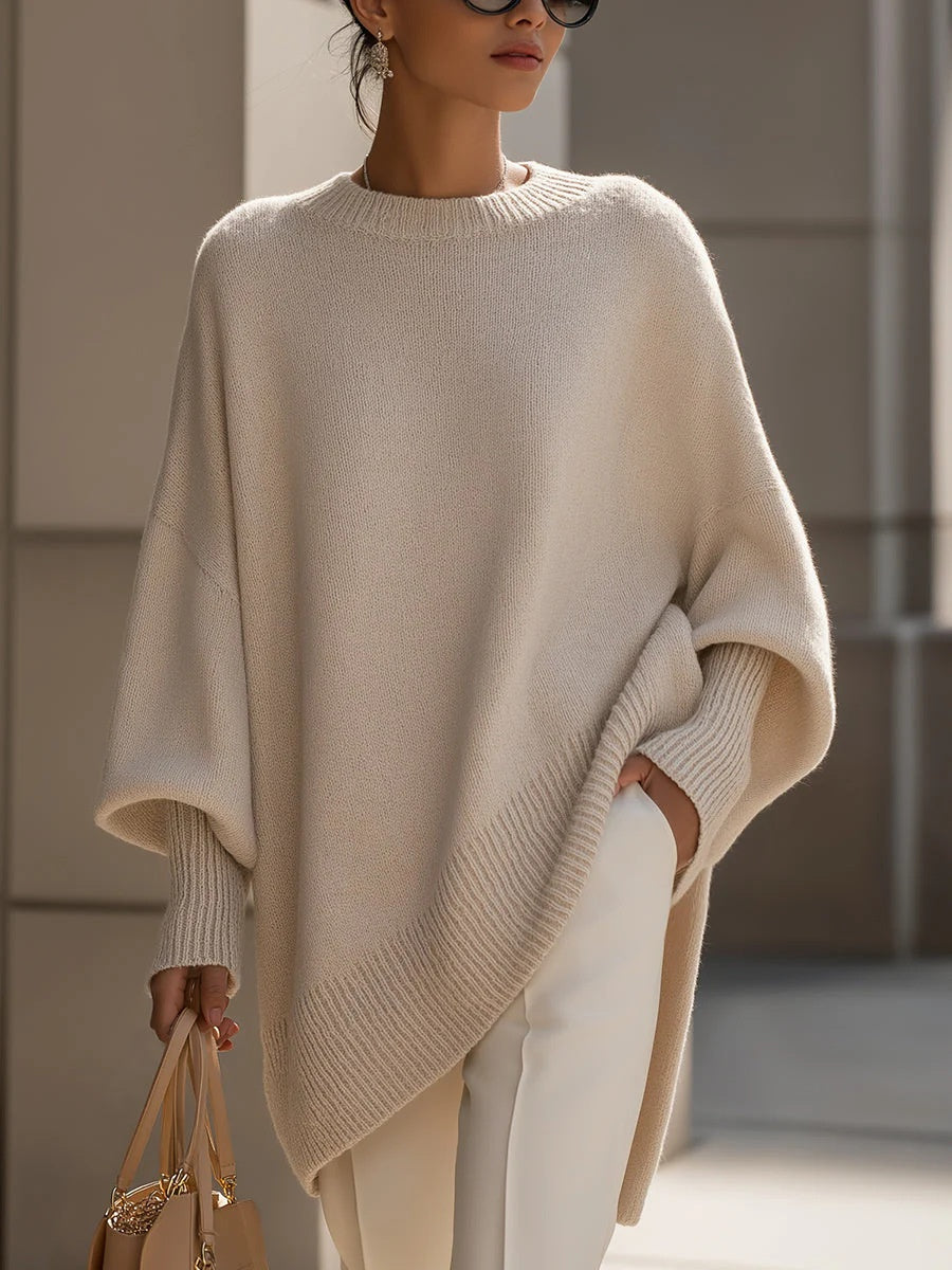 A woman wearing a cream-colored oversized knit sweater with wide batwing sleeves and a ribbed hem, paired with white pants and holding a beige handbag.