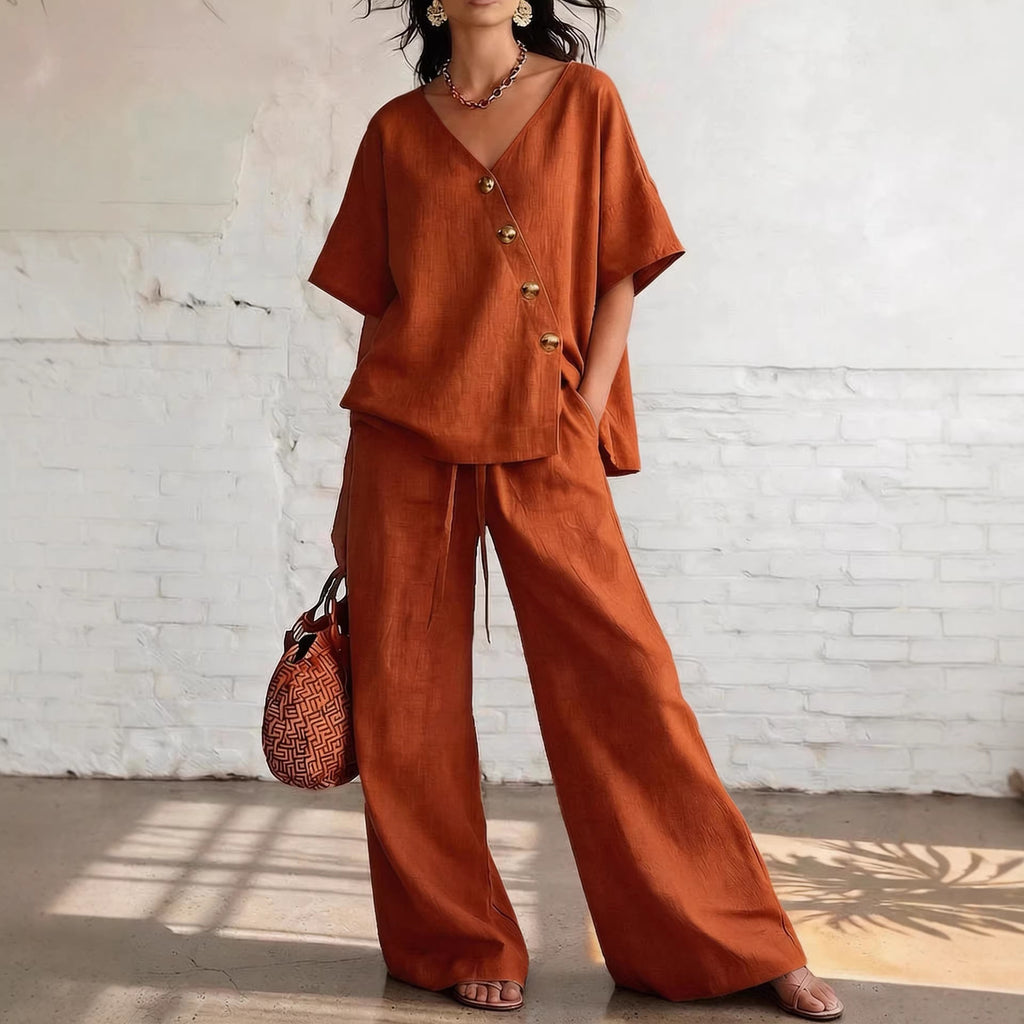 Woman wearing a rust-colored outfit with wide-leg pants and a top, holding a matching bag against a white brick wall.