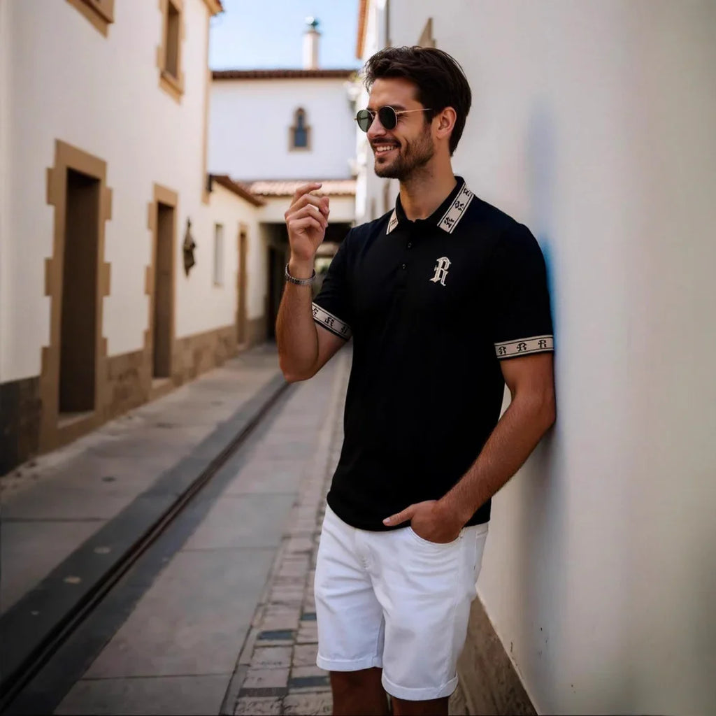 Man wearing a black polo shirt with a logo and white shorts standing in a narrow street.