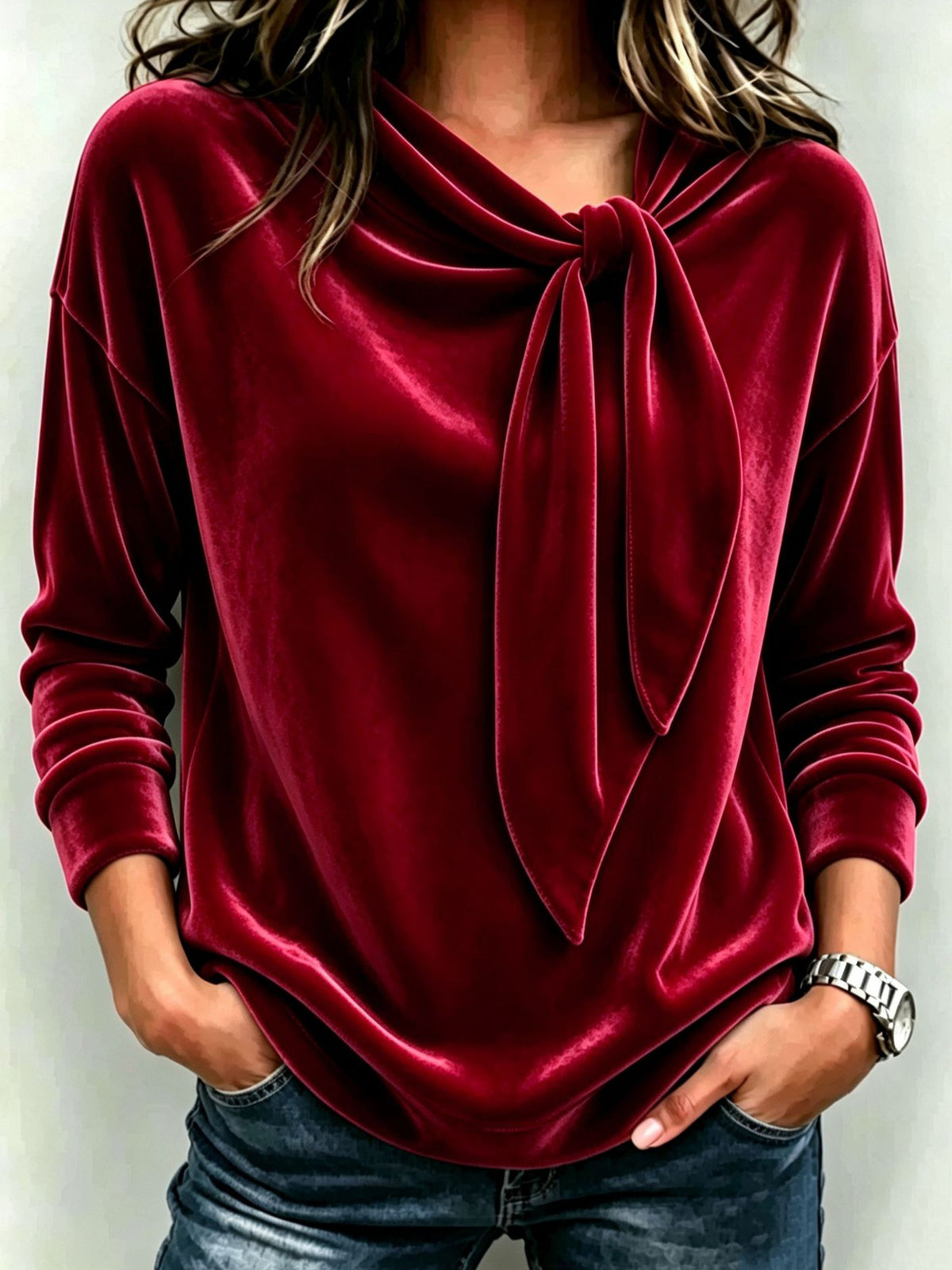 A woman wearing a long-sleeve velvet top in deep burgundy red with a cowl neck and front tie detail, paired with blue jeans.