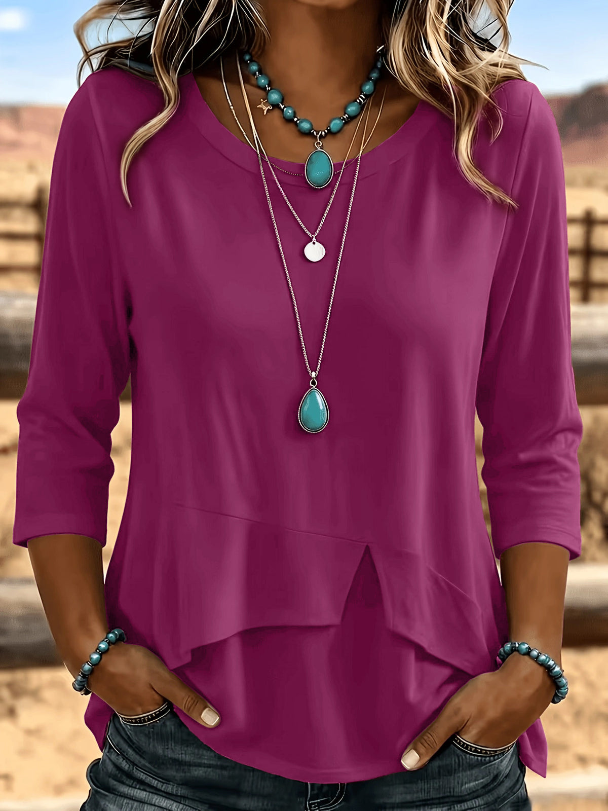 A woman wearing a magenta three-quarter sleeve top with a layered high-low hem, styled with layered turquoise and silver necklaces and blue jeans, set against a blurred outdoor desert background.