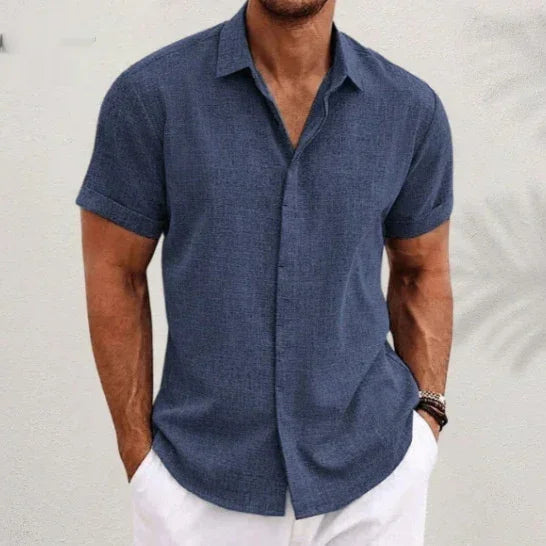 Man wearing a blue short-sleeve shirt and white pants against a light background