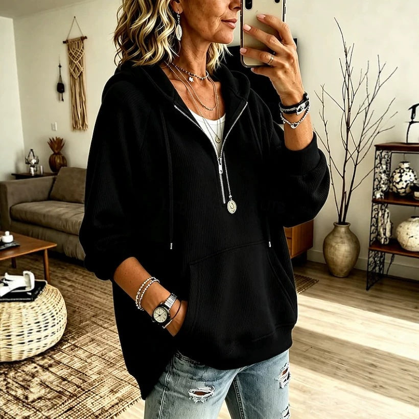 A woman takes a mirror selfie in a living room while wearing a black zip-up hoodie with a front kangaroo pocket, layered over a white top and light-wash distressed jeans.
