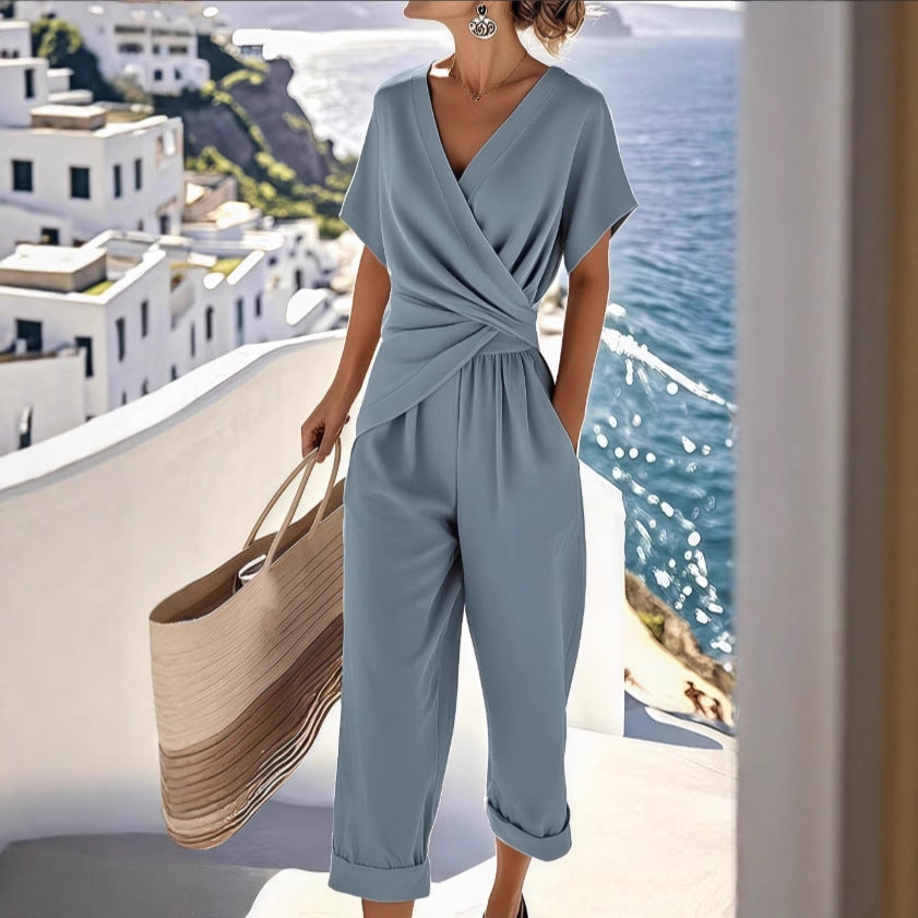 Woman in a light blue jumpsuit holding a straw bag with a scenic background of buildings and ocean.