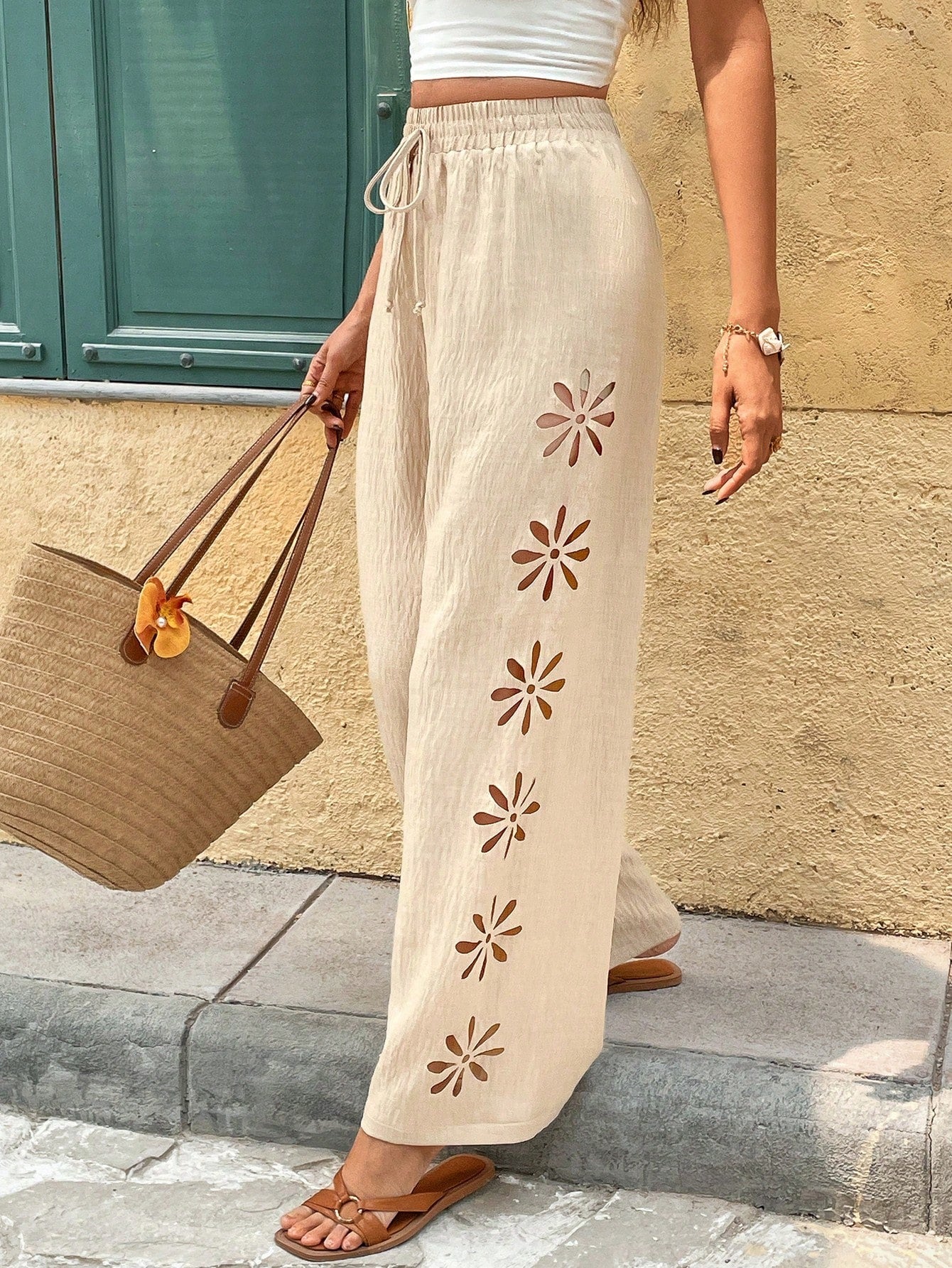 Person wearing beige pants with floral cutouts, holding a straw bag against a wall.