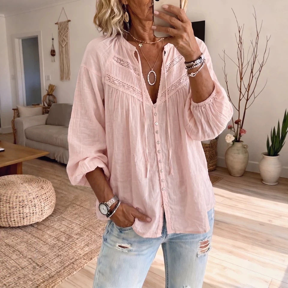 A woman takes a mirror selfie in a bright living room, wearing a light pink long-sleeve button-up blouse with lace detailing at the neckline and light-wash distressed jeans.
