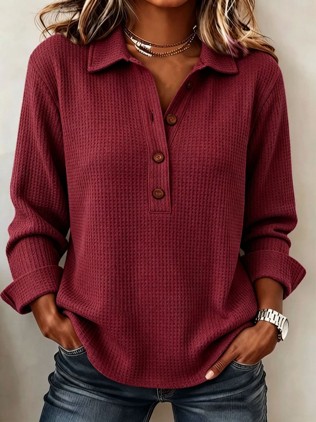 A woman wearing a long-sleeve waffle-knit polo shirt in burgundy with brown buttons, paired with blue jeans and layered necklaces.