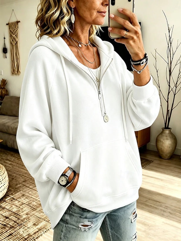 A woman takes a mirror selfie wearing a white zip-up hoodie with drawstrings and a front kangaroo pocket, paired with distressed light-wash jeans. The setting is a cozy, modern living room with a couch, woven rug, and a potted plant in the background.
