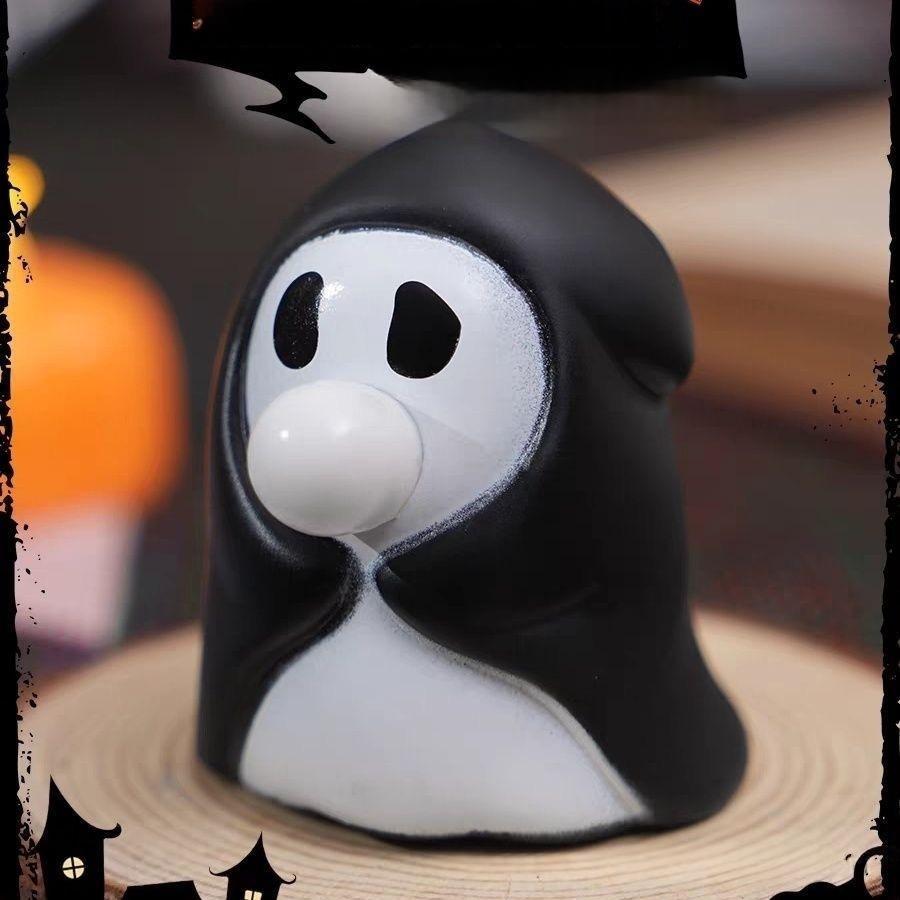 A black and white ghost-shaped novelty item, possibly a squishy toy or decorative figurine, shown close-up on a wooden surface with a blurred Halloween-themed background.