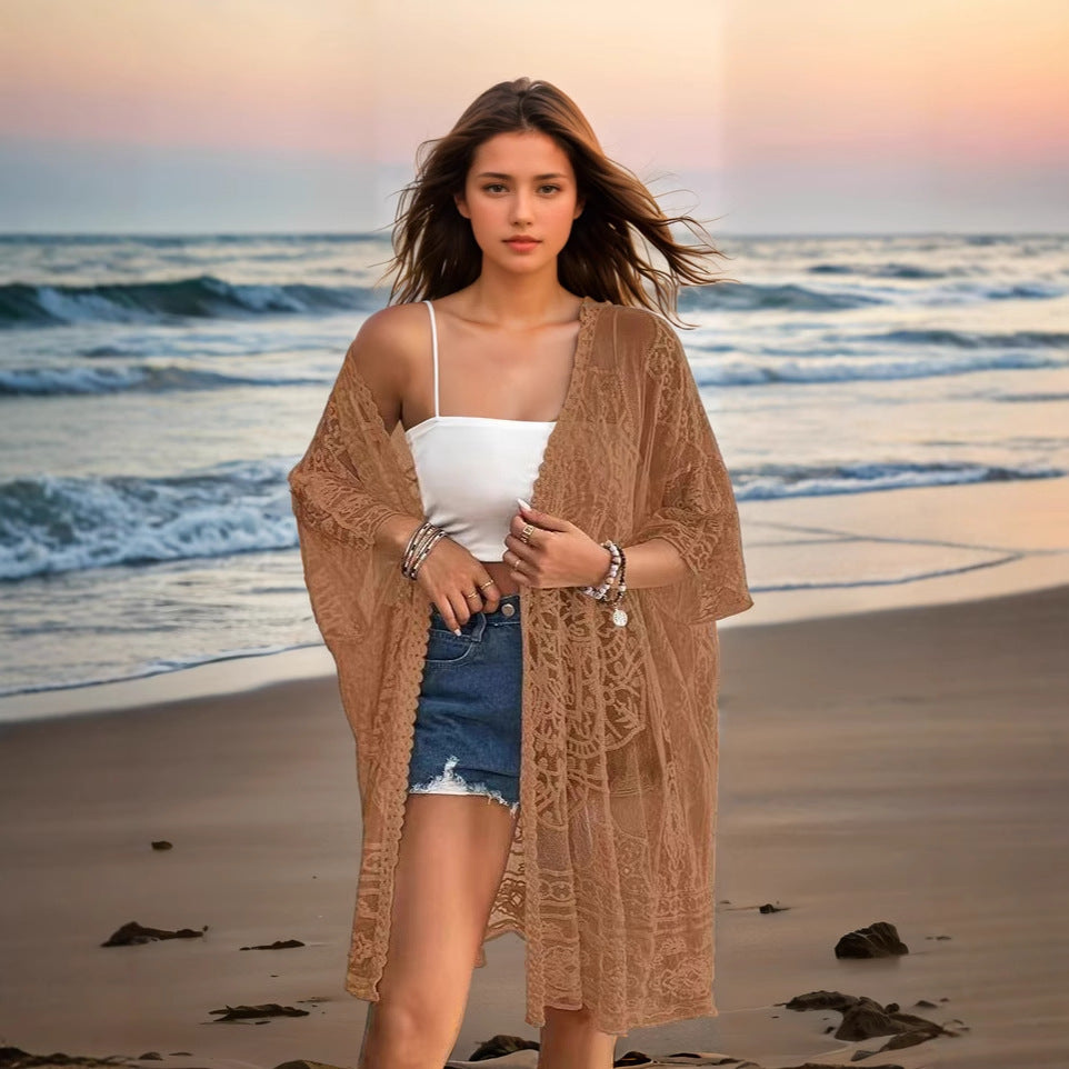 Woman standing on a beach wearing a beige lace cover-up, white top, and denim shorts.