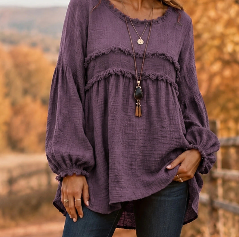 A woman wearing a loose, long-sleeve purple top with ruffled trim and gathered cuffs, paired with dark blue jeans, standing outdoors against a blurred autumn landscape.
