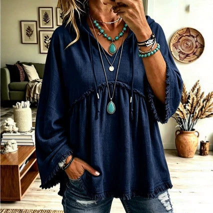 A woman wearing a loose navy blue bohemian-style tunic top with fringed edges, layered necklaces, and turquoise jewelry, standing in a decorated living room.