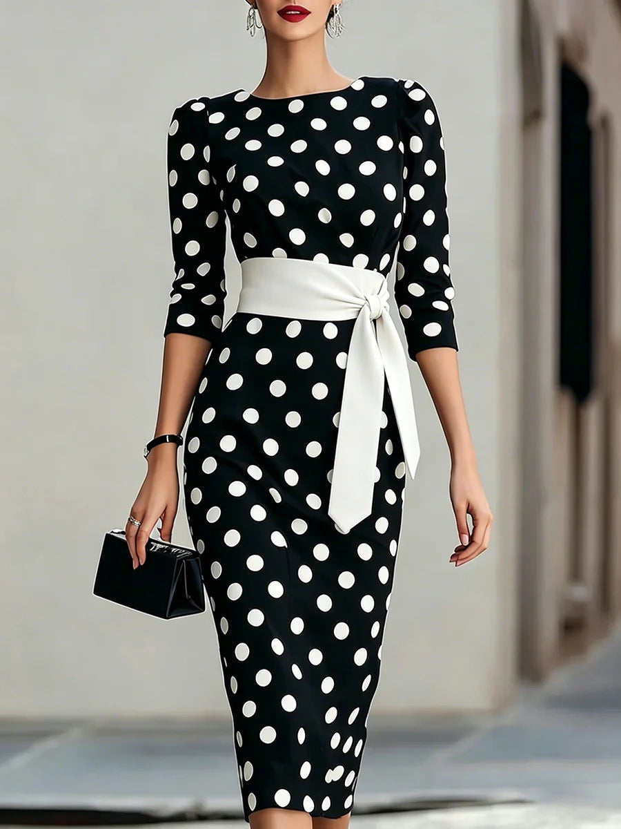 A woman models a black and white polka dot midi dress with three-quarter sleeves and a wide white waist sash tied in a bow. She is holding a small black clutch against a blurred outdoor background.