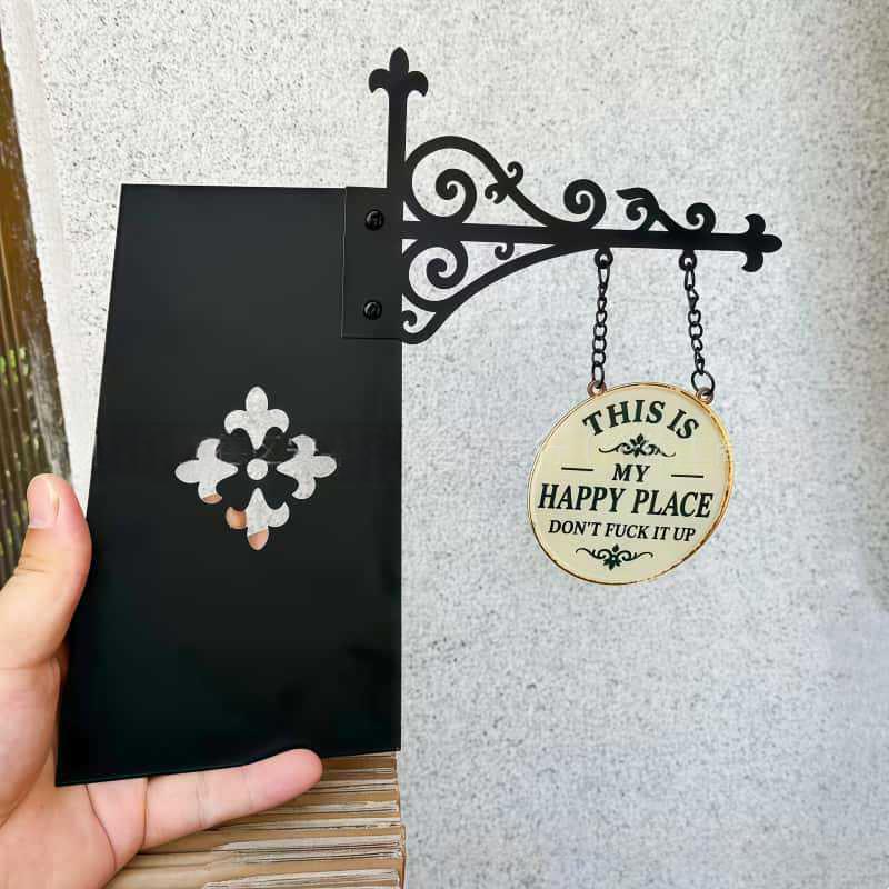 A hand holds a black metal sign holder against a textured gray wall. The holder features an ornate scrollwork bracket with a hanging chain and round wooden sign that reads, 'THIS IS MY HAPPY PLACE DON'T FUCK IT UP.
