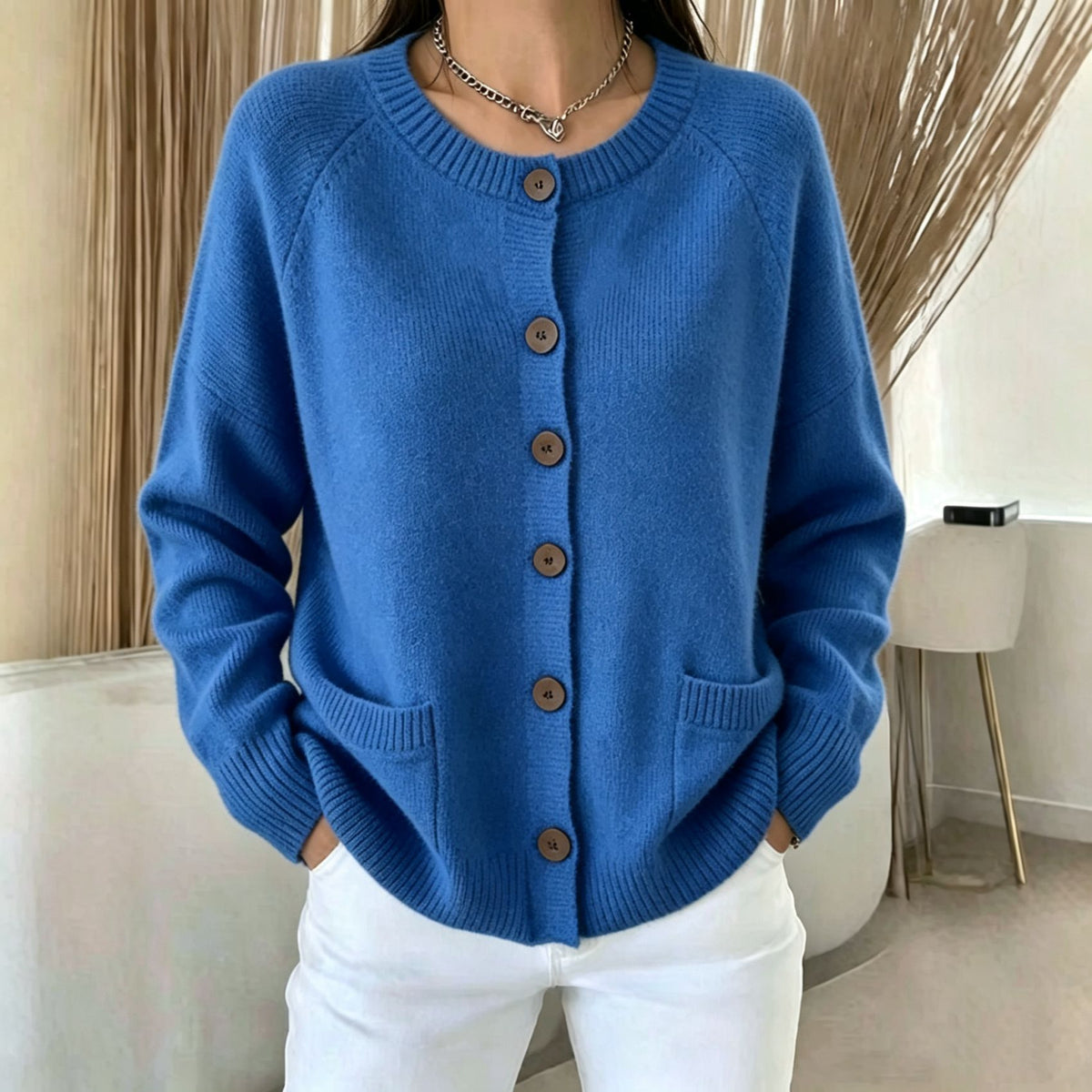 A woman wearing a blue knit cardigan sweater with brown buttons and white pants, standing indoors in a modern room with neutral decor.