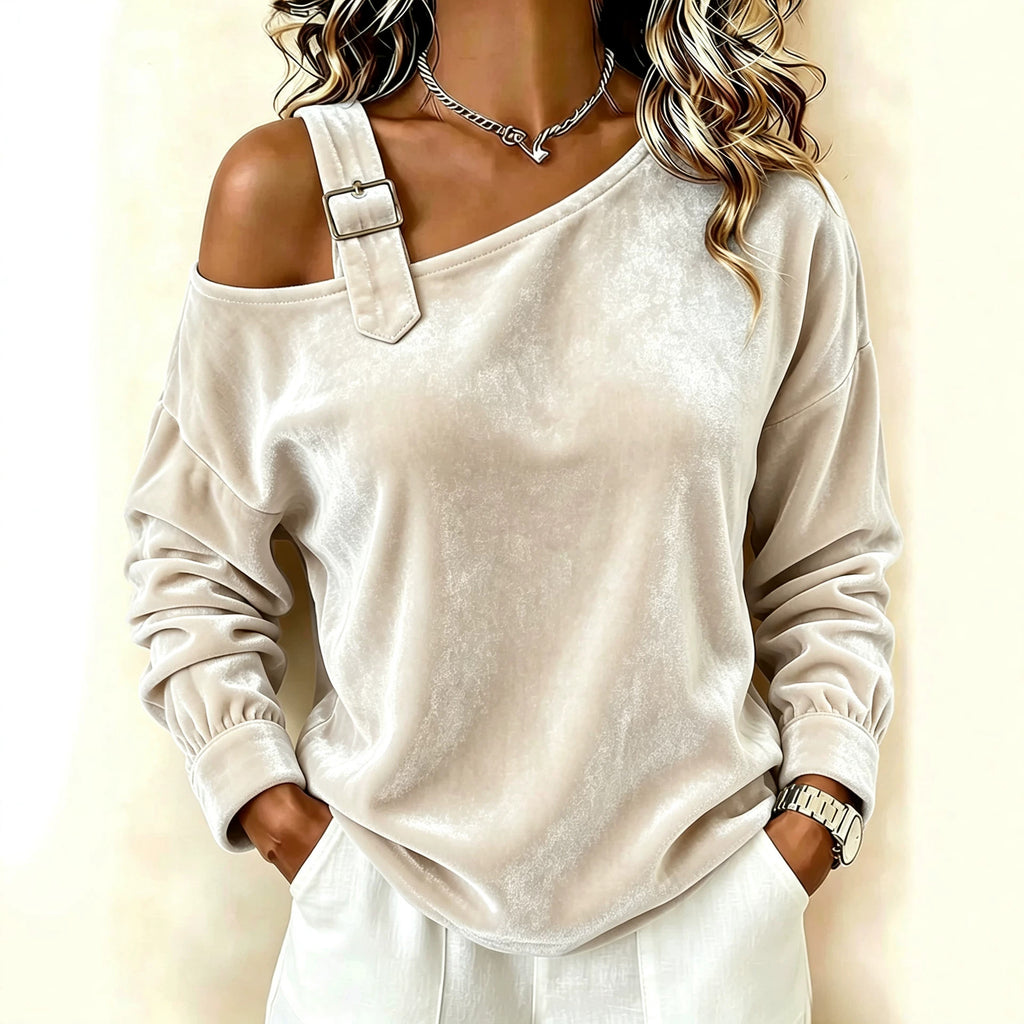 A woman wearing a cream-colored off-the-shoulder velvet top with a decorative strap and buckle detail, paired with white pants, set against a soft beige background.