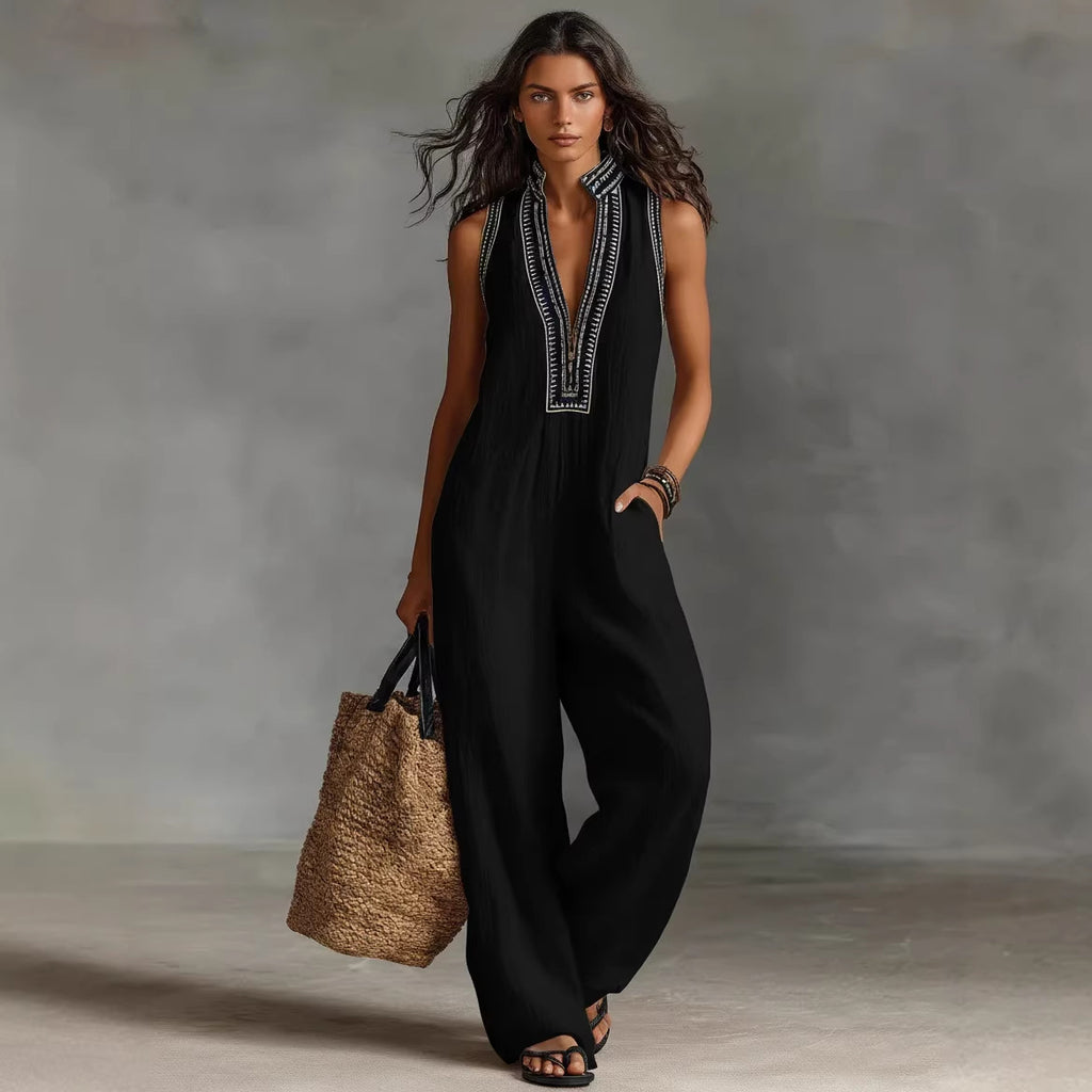 Woman wearing a black sleeveless jumpsuit with white embroidery, holding a woven bag against a gray background.