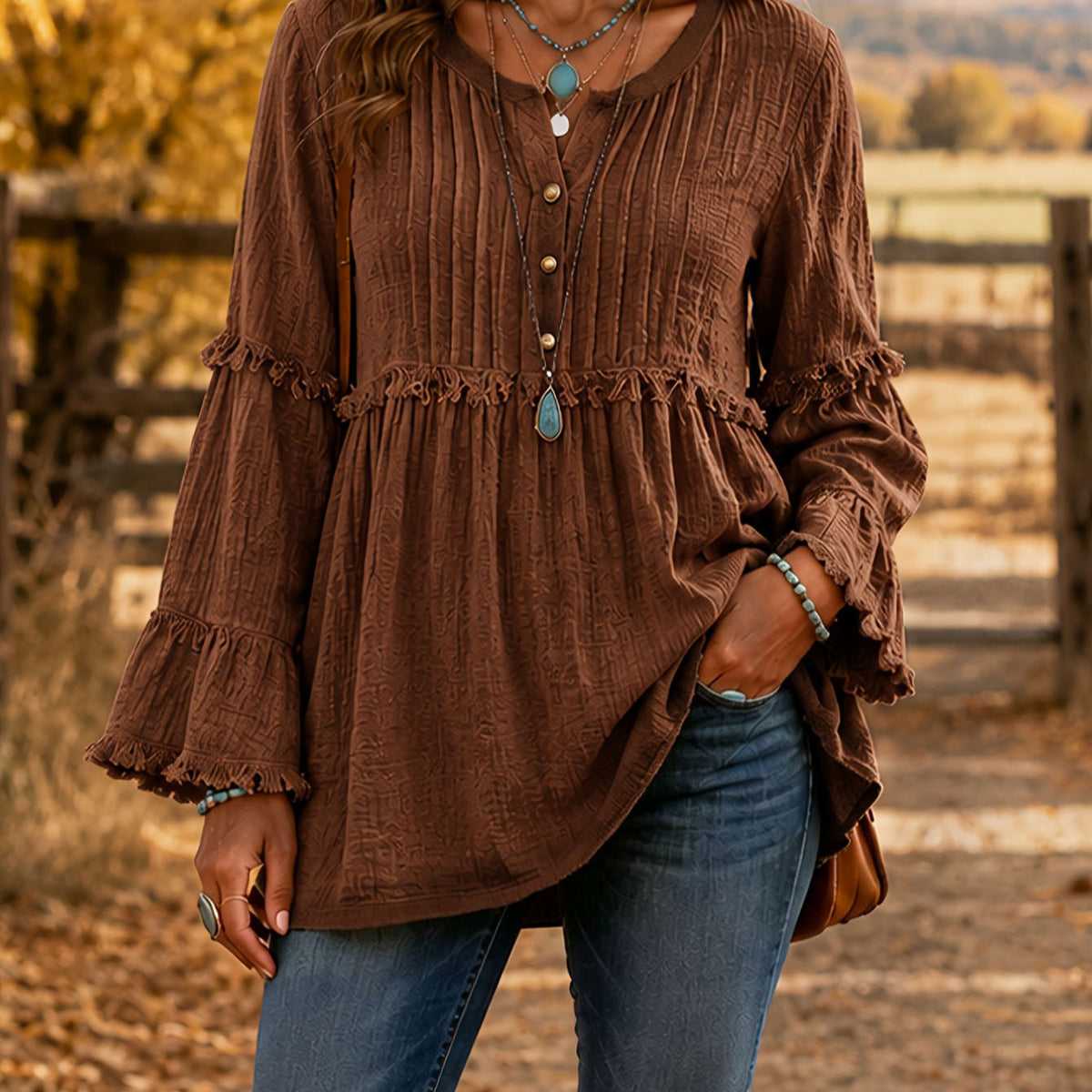 A woman wearing a brown long-sleeve crinkle-textured blouse with ruffled trim and tiered bell sleeves, paired with blue jeans, standing outdoors in front of a rustic wooden fence.