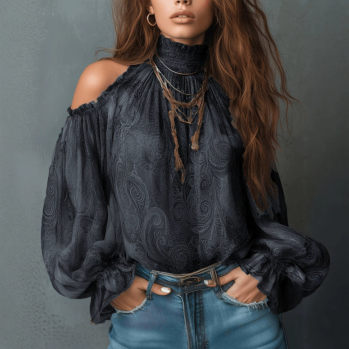 A woman wearing a dark charcoal gray cold-shoulder blouse with a subtle paisley pattern, paired with blue jeans and layered gold necklaces, set against a textured gray background.